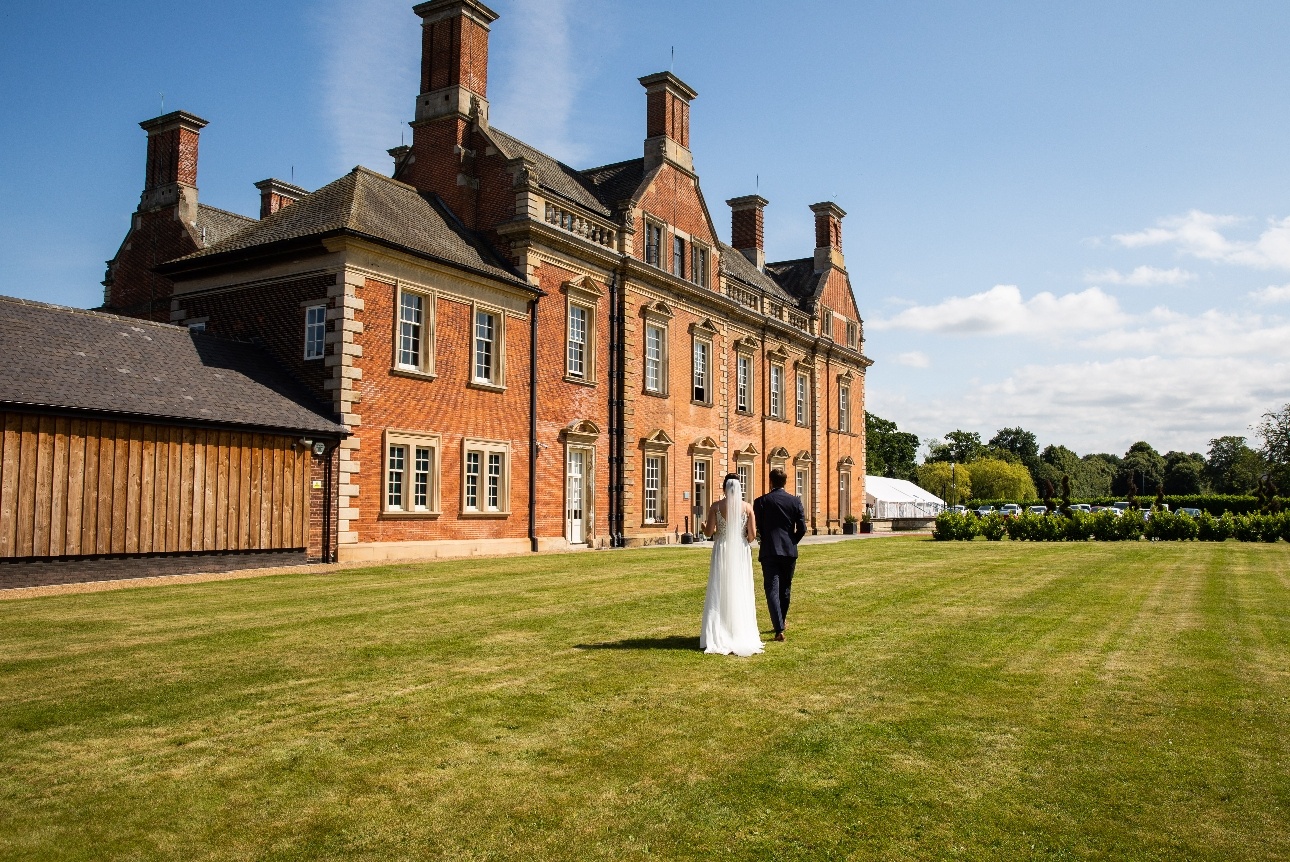 red brick manor house with manicured grass and a bride and groom walking across it