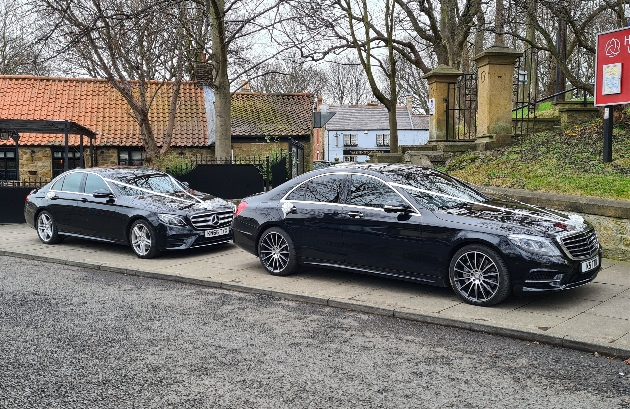 two black saloon cars parked in a street with wedding ribbons on 