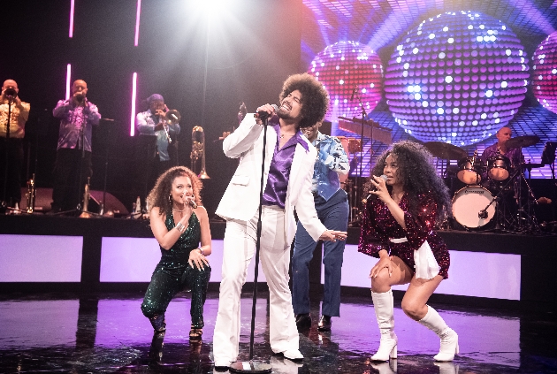 male singer in white suit on stage with two female dancers and singer in glittery outfits