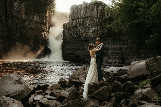 A bride and groom embracing next to a river with a waterfall in the background