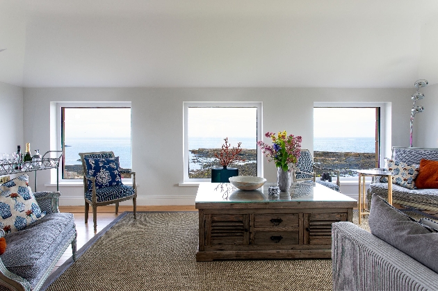 A living room with grey sofas, a coffee table in the centre and windows looking out to the sea
