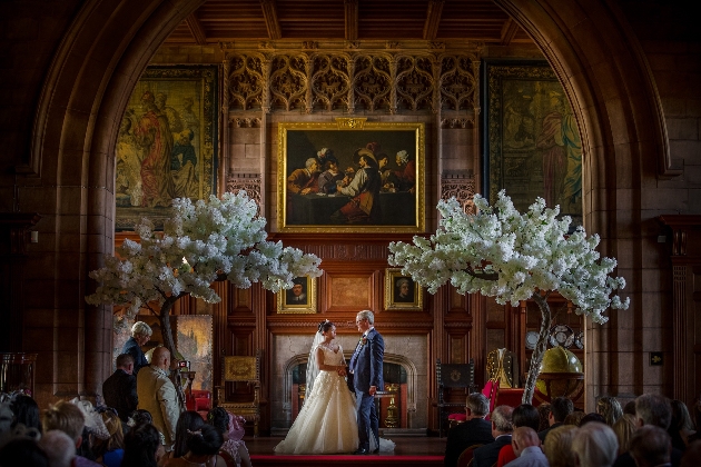 A bride and groom standing at the end of the alter surrounded by grand paintings and floral trees