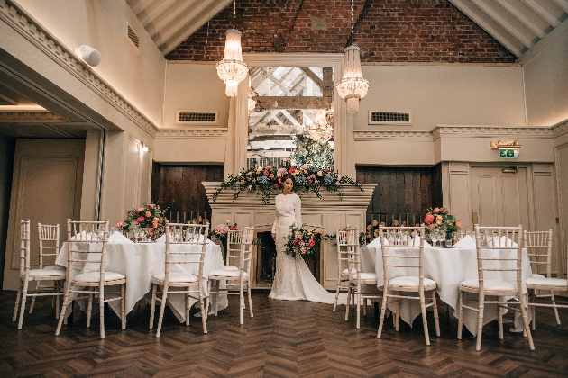 A bride standing in a grand room with a fireplace, large mirror and two round tables decorated with flowers
