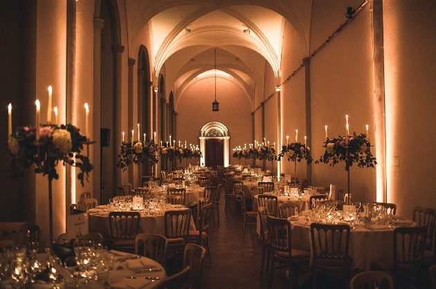 A dark lit room with high ceilings and lots of round tables decorated with flowers