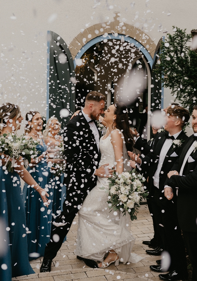 A bride and groom kissing while their friends throw confetti over them