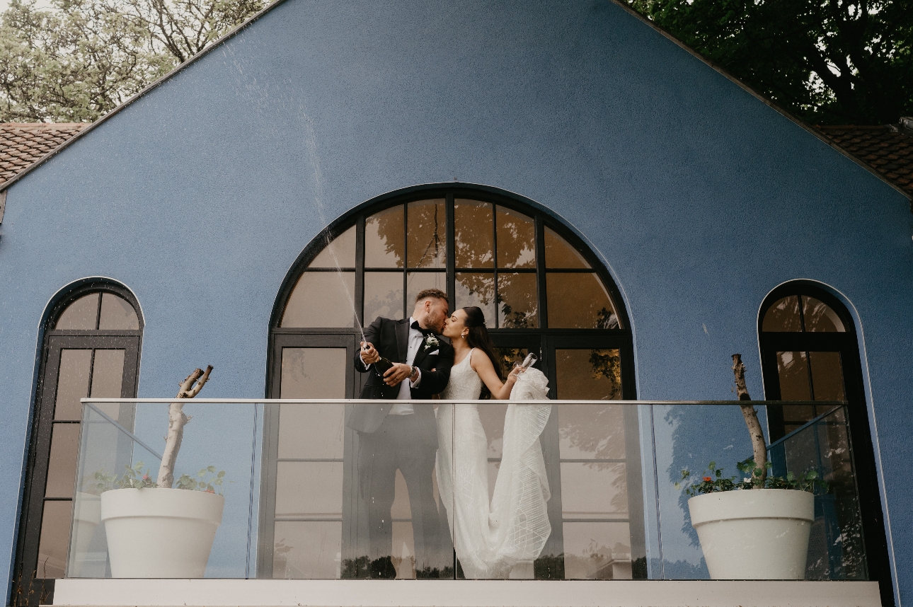 A bride and groom holding a bottle of drink in front of a blue building